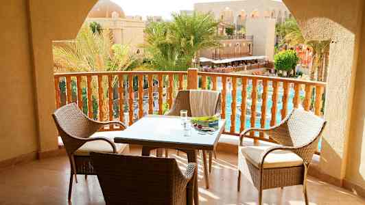 Udsigt fra en balkon med bord og stole samt udsigt over en pool og palmer ved en arkitektonisk bygning. (AI-genereret tekst)