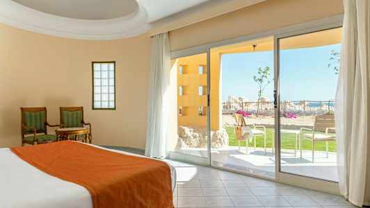 Værelse med seng og sengetøj i orange, to stole med et bord og udsigt til en terrasse med grønne områder og liggestole. (AI-genereret tekst)