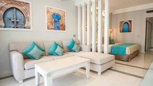 Et opholdsrum med en beige sofa og turkise puder, et hvidt sofabord og kunstværker på væggen, delvist adskilt med søjler fra et soveområde med en seng. (AI-genereret tekst)