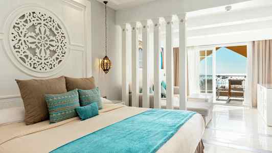 Et hotelværelse med lyse vægge, en dekoreret seng med blå og beige sengetøj, samt en altan med havudsigt. (AI-genereret tekst)