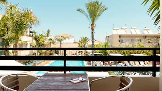 Udsigt fra en balkon med bord og stole mod en pool med liggestole og omkringliggende palmer. (AI-genereret tekst)