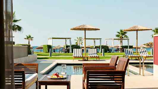 Udendørsmøbler ved en pool med liggestole og parasoller, græsplæne og hav i baggrunden. (AI-genereret tekst)