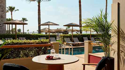 Terrasse med bord, stole, udsigt over poolen og liggestole under parasoller. (AI-genereret tekst)