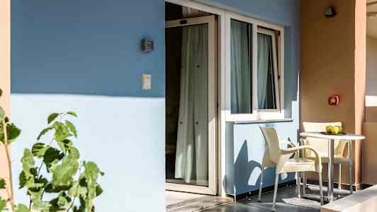 En balkon med to stole og et bord står foran en bygning med blå og beige vægge. (AI-genereret tekst)