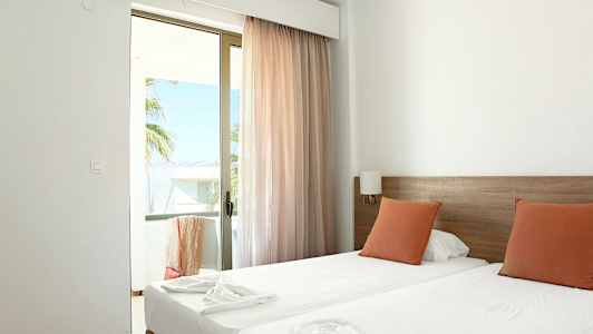 Et hotelværelse med to enkeltsenge, orange puder og en glasdør, der fører til en altan. (AI-genereret tekst)