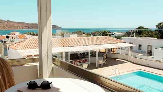 Udsigt fra en balkon over en pool, dele af hotelområdet og havet i baggrunden. (AI-genereret tekst)