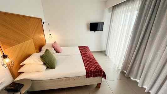 Billede af et hotelværelse med en dobbeltseng, et træpanel som sengegavl, røde og grønne pyntepuder, hængende lamper, en lille bordtelefon og gardiner. Et TV hænger på væggen. (AI-genereret tekst)