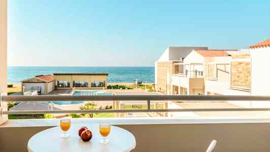 Udsigt fra en altan med bord, hvor to glas og to æbler står, mod en pool, bygninger og havet. (AI-genereret tekst)