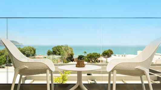 Udsigt fra en altan med to hvide stole og et bord med en lille plante samt havet og grønne områder i baggrunden. (AI-genereret tekst)