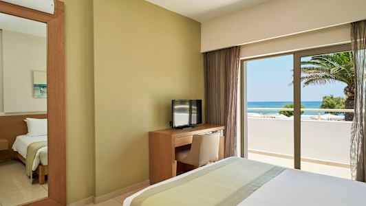 Hotelværelse med seng, fjernsyn og skrivebord samt udsigt over havet gennem et vindue. (AI-genereret tekst)