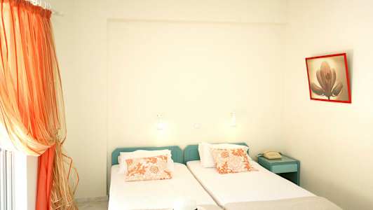 To enkeltsenge med orange mønstrede puder, en telefon på et blåt natbord og orange gardiner ved vinduet. (AI-genereret tekst)