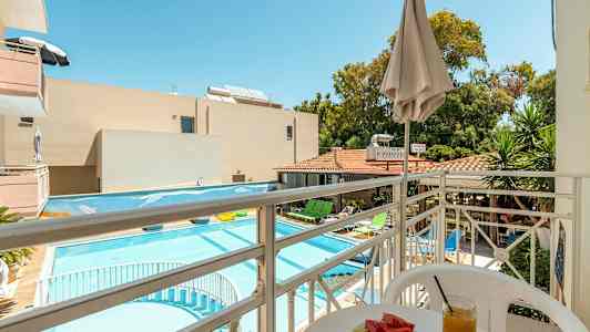 Udsigt fra en balkon mod en pool og hotelområde med liggestole, parasoller og grønne områder. På balkonen står et bord med en tallerken melon og et glas juice. (AI-genereret tekst)