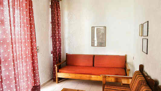 Stuen har røde gardiner, en træsofa med røde ternede puder og en stofbetrukket stol ved siden af. (AI-genereret tekst)