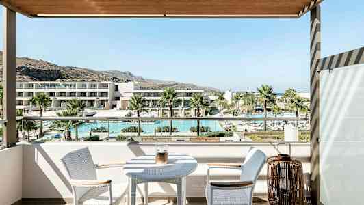 Udsigt fra en balkon med et bord og to stole, der viser en pool, palmer og moderne bygninger i baggrunden. (AI-genereret tekst)