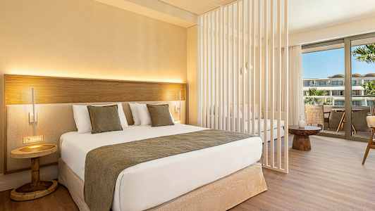 Et moderne hotelværelse med en dobbeltseng, dekorative puder og et lille natbord. Værelset har en skillevæg og et siddeområde med udsigt til en balkon. (AI-genereret tekst)