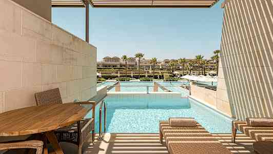 En terrasse med liggestole ved siden af en pool. I baggrunden ses flere bygninger og palmer. (AI-genereret tekst)