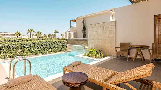 Billedet viser en terrasse med liggestole, et lille bord og to stole ved siden af en privat pool. I baggrunden ses frodige buske og palmer. (AI-genereret tekst)