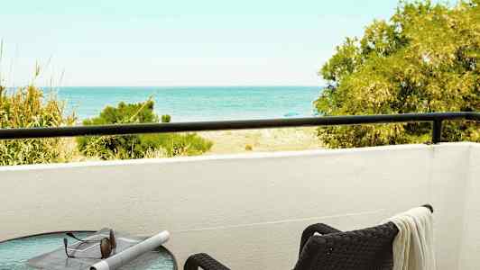 En altan med udsigt til havet, et bord med solbriller og en avis samt en stol med et håndklæde. (AI-genereret tekst)