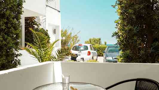 Terrasse med et bord, hvor en bog og et glas står. Udenfor ses parkerede biler og vegetation. (AI-genereret tekst)