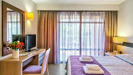 Et hotelværelse med en seng dækket med et lilla sengetæppe, skrivebord med et fjernsyn og en blomsterbuket samt et stort vindue med gennemsigtige gardiner. (AI-genereret tekst)