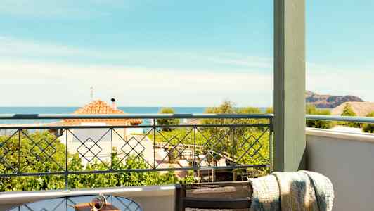 Udsigt fra en balkon med et glasbord og stol. Havet og huse ses i baggrunden. (AI-genereret tekst)