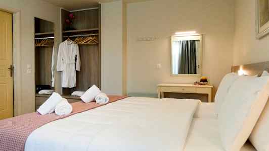 Et hotelværelse med en dobbeltseng, sengetøj i hvidt og et rosa tæppe. Et skab med hængende tøj og et spejl ses i baggrunden. (AI-genereret tekst)