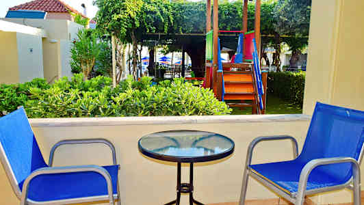 To blå stole ved et bord med glasbordplade på en terrasse, med legeplads og grønne områder i baggrunden. (AI-genereret tekst)