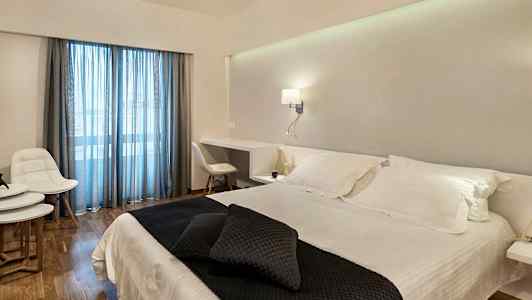 Et hotelværelse med en dobbeltseng med hvide lagner og mørke pyntepuder. Værelset har et skrivebord, hvide møbler og glasdøre med blå gardiner. (AI-genereret tekst)