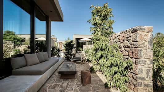 Stenbelagt terrasse med sofa, bord og stole nær et glashus, omgivet af planter og mure. (AI-genereret tekst)