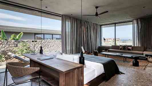 Moderne hotelsuite med stor seng, skrivebord og siddeområde. Vinduer med udsigt over havet og grønne omgivelser. (AI-genereret tekst)