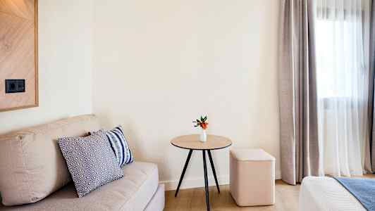 Et værelse med en beige sofa, et træbord med en lille hvid vase og orange blomster, en blød skammel og gardiner ved en vinduesudskæring. (AI-genereret tekst)