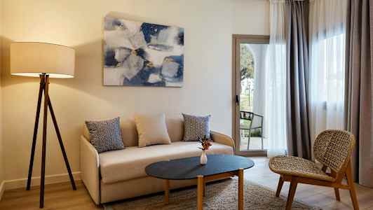 En stue med en sofa, et bord og en stol samt en gulvlampe og gardiner foran vinduet. (AI-genereret tekst)