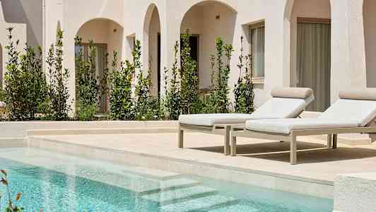 Billede af to solsenge ved en pool, med hvide bygninger og grønne planter i baggrunden. (AI-genereret tekst)