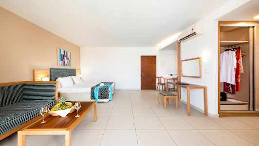 Et rummeligt hotelværelse med dobbeltseng, siddeområde, spisebord, spejl og garderobe. En frugtskål og vinglas står på sofabordet. (AI-genereret tekst)