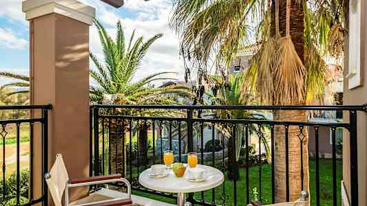 Udsigt fra en balkon med bord, stole, drikkevarer og frugt samt palmer og have i baggrunden. (AI-genereret tekst)