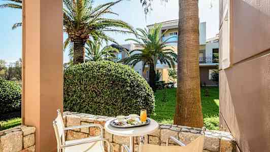 Udsigt fra en terrasse med bord og stole mod en frodig have med palmer og flerboligbygninger. (AI-genereret tekst)