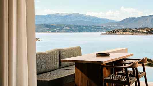 Udsigt over et træbord og stole med havet og bjerge i baggrunden. (AI-genereret tekst)