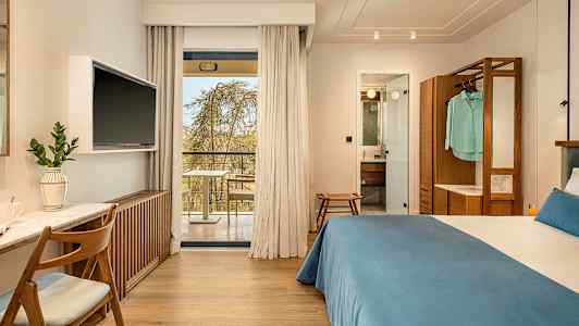 Hotelværelse med dobbeltseng, skrivebord og TV. Glasdøre fører ud til en balkon. Badeværelse synligt gennem en åben dør. (AI-genereret tekst)