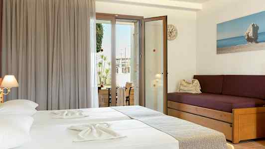 Et hotelværelse med to senge med lyse lagner, en lilla sofa med puder og en glasdør, der fører ud til en balkon. (AI-genereret tekst)