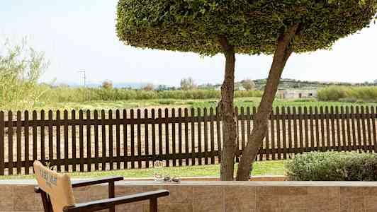 Udsigt fra en terrasse mod et træhegn og et grønt landskab. (AI-genereret tekst)