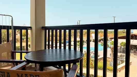 Udsigten fra en altan med bord og stole med en pool og omkringliggende bygninger i baggrunden. (AI-genereret tekst)