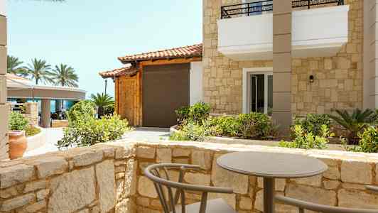 En terrasse med et bord og stole omgivet af stenmur, med udsigt til en bygning med stenfacade og palmer i baggrunden. (AI-genereret tekst)