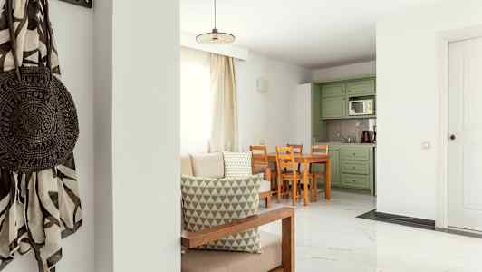 Et opholdsstue med lys indretning, bord og stole samt grønne køkkenskabe. (AI-genereret tekst)