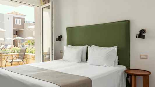 Hotelværelse med en seng og en grøn sengegavl, lyse vægge og vindue med udsigt over en terrasse. (AI-genereret tekst)