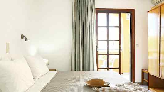 Et soveværelse med en seng, puder og ét tæppe. Et vindue med gardiner og et natbord er synligt. (AI-genereret tekst)