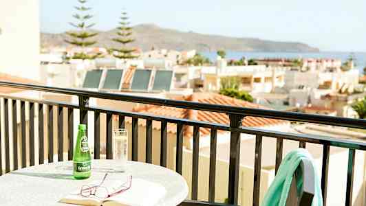 Bord med bog, briller og drikkevare på altan med udsigt over huse og hav. (AI-genereret tekst)