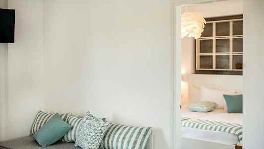 Et hotelværelse med en sofa med puder i forgrunden og en redt seng i baggrunden. (AI-genereret tekst)