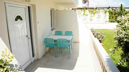 Billede af en terrasse med et bord og fire turkise stole, omgivet af grønne områder. (AI-genereret tekst)