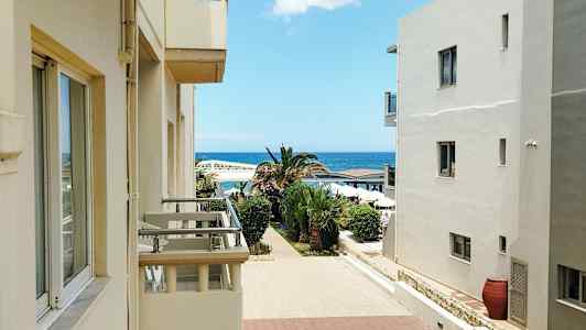 Bygninger med altaner åbner op mod en gangsti omgivet af palmer, med havet i baggrunden. (AI-genereret tekst)