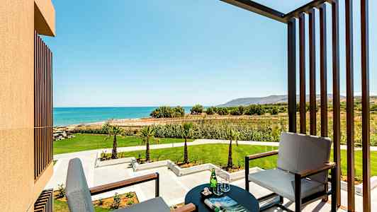 Billede af en altan med to siddepladser, et bord med glas og avis, med udsigt over en grøn have og blå hav. (AI-genereret tekst)